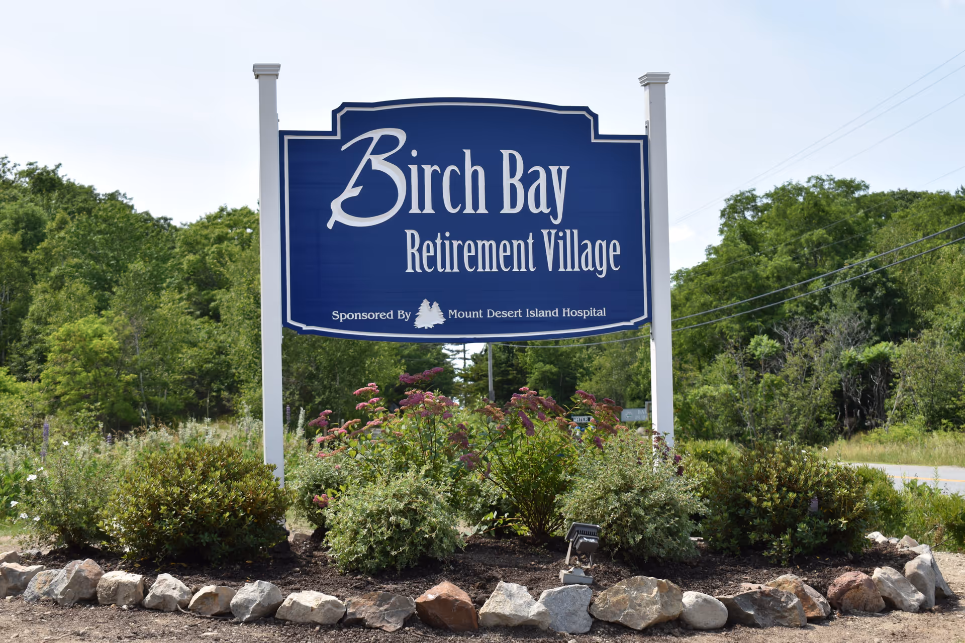A large blue sign with white text reading 'Birch Bay Retirement Village' sponsored by Mount Desert Island Hospital, surrounded by green bushes and flowers with a background of trees and a clear sky.