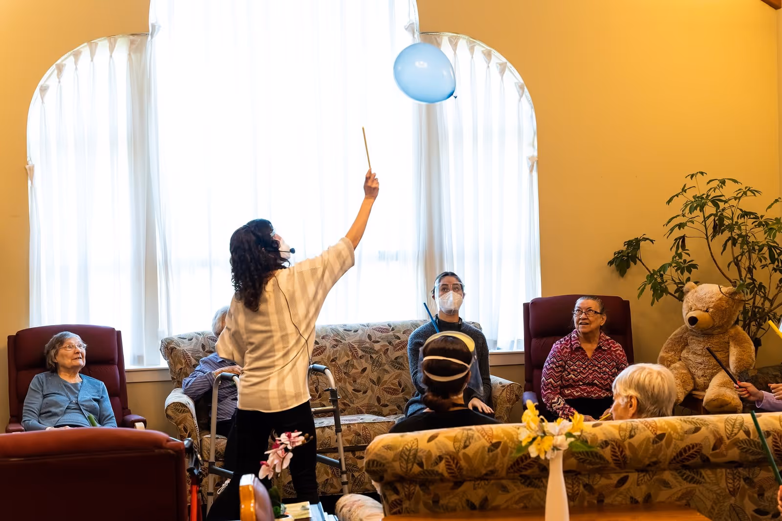 A group of elderly people sitting in a living room area with a caregiver standing and holding a stick to keep a blue balloon in the air. The room has large windows with white curtains, comfortable chairs, a sofa, a large teddy bear, and a potted plant.