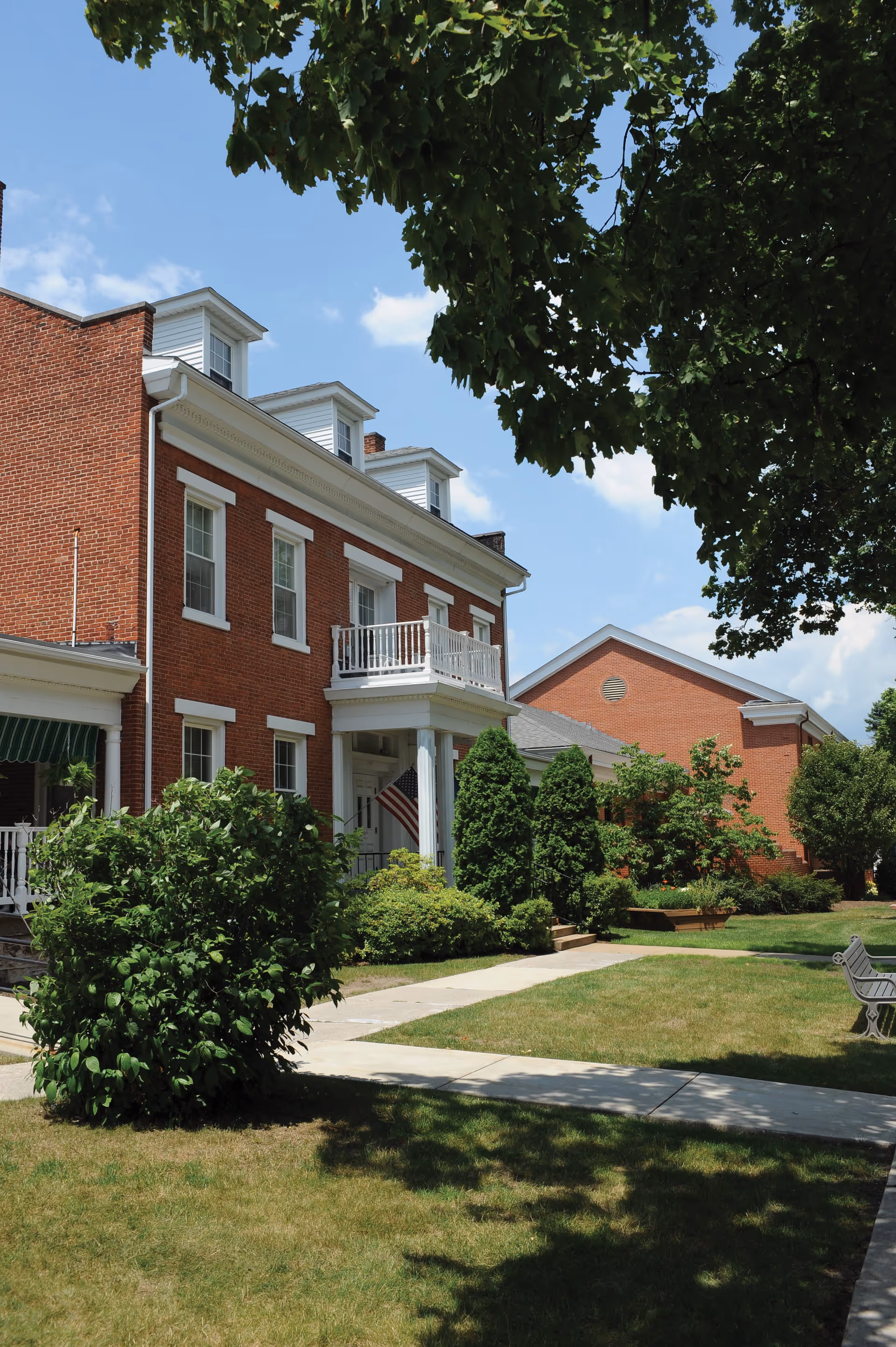 Exterior view of a red brick building with white trim and a small balcony above the entrance. The building is surrounded by green bushes, trees, and a well-maintained lawn with a concrete walkway and a bench. The sky is clear with a few clouds.