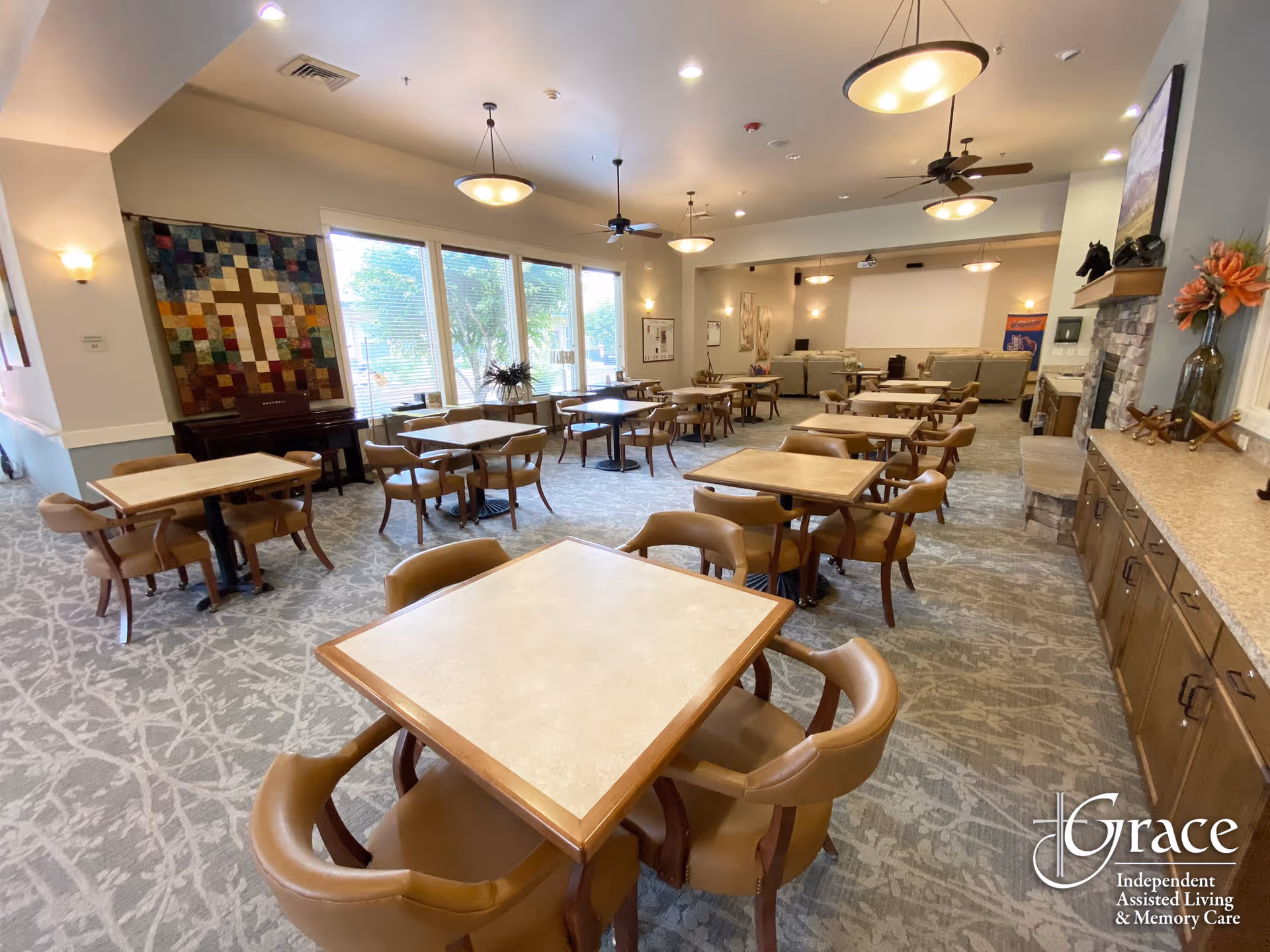 A spacious dining area with multiple square tables and brown cushioned chairs arranged neatly. Large windows on one side let in natural light, and the room features a carpeted floor with a patterned design. There is a colorful quilt with a cross hanging on the wall, ceiling fans, pendant lights, and a fireplace with decorative items on the mantel. The room appears clean and inviting, designed for communal dining and socializing.