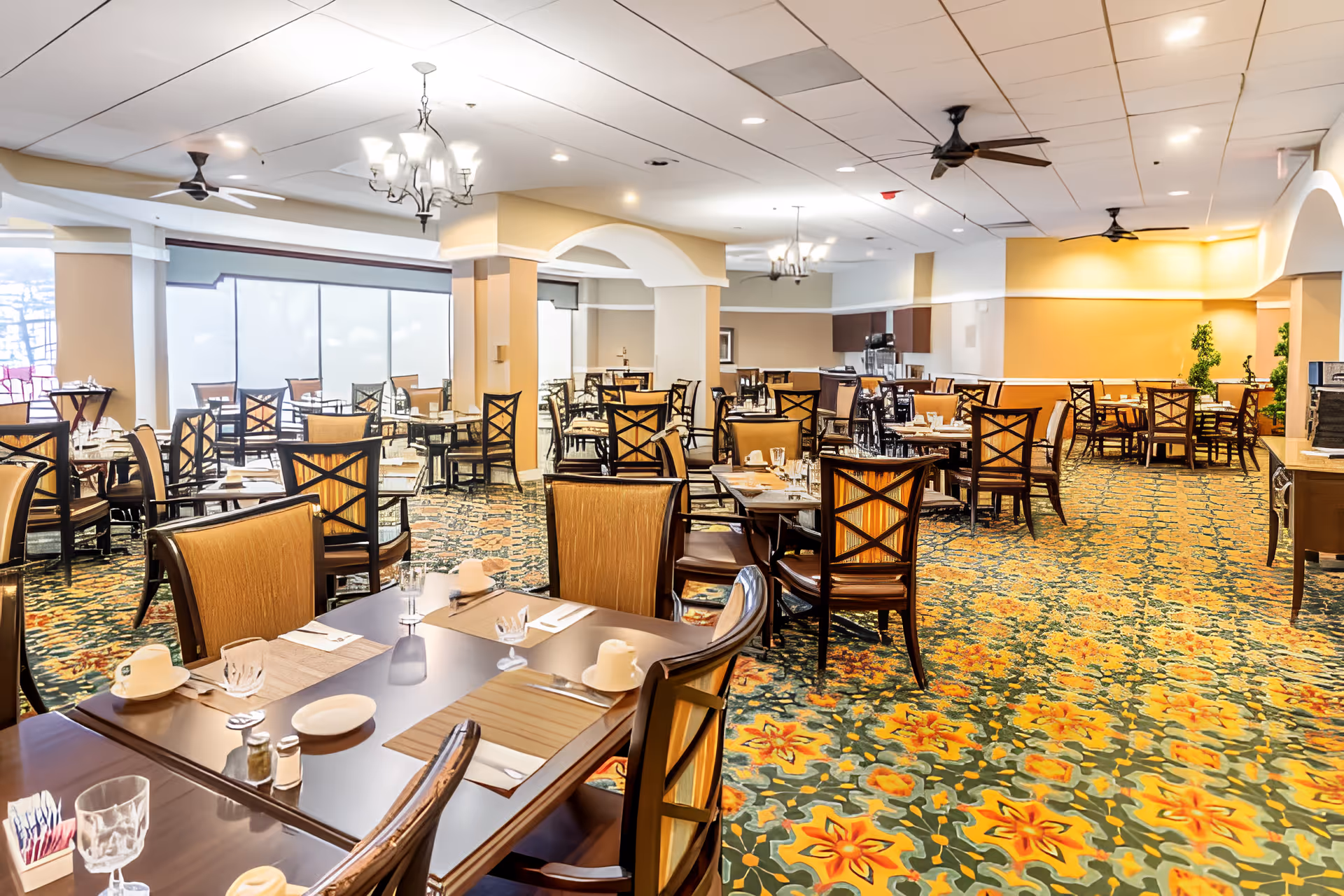 A spacious dining room with multiple wooden tables and chairs arranged neatly. The tables are set with placemats, cups, glasses, and utensils. The room features a colorful floral carpet, ceiling fans, chandeliers, and large windows with frosted glass panels allowing natural light to enter. The walls are painted in warm tones with white trim and arches.