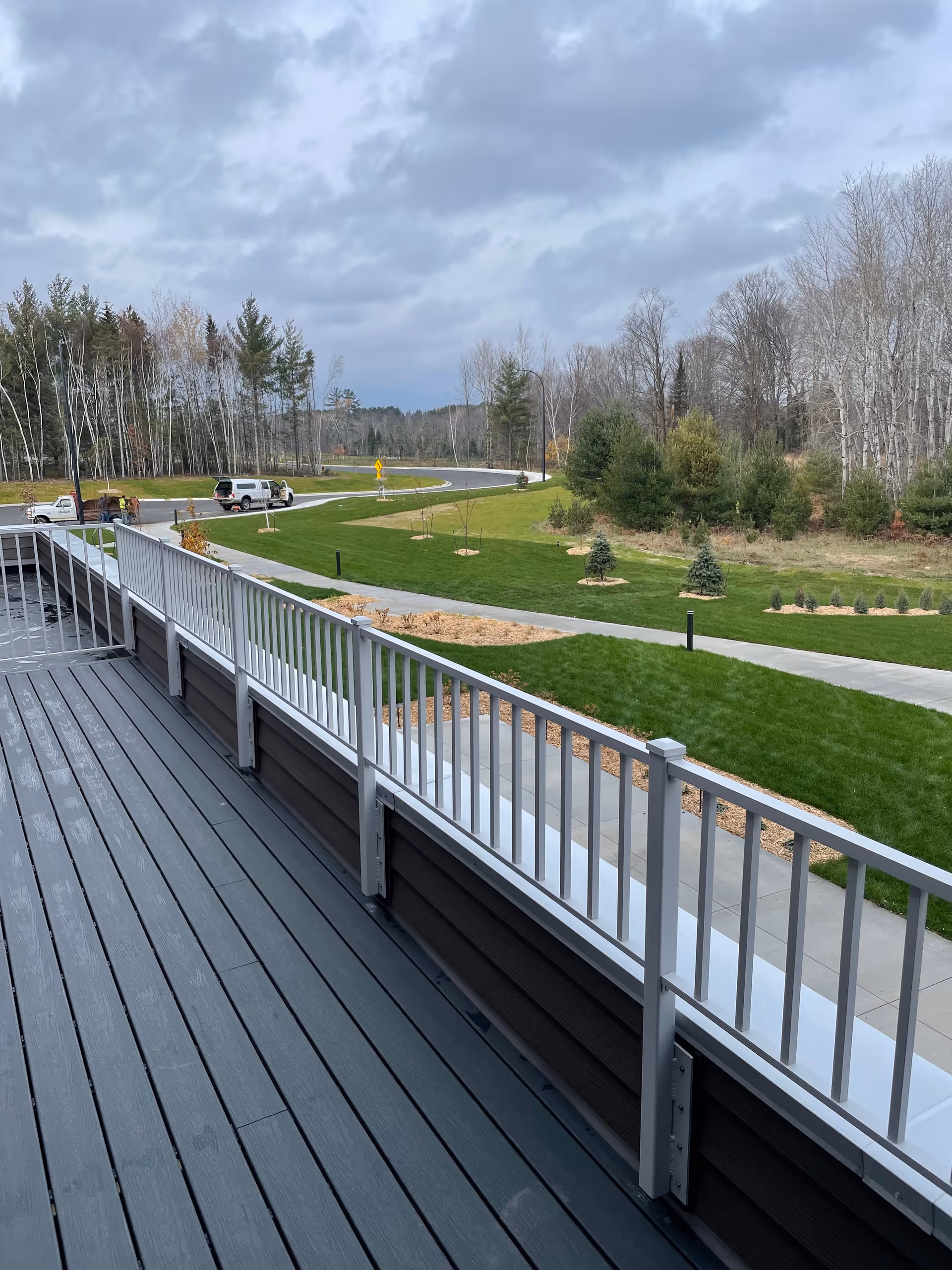 View from a balcony with gray wooden flooring and white railing overlooking a landscaped area with green grass, small trees, a curved road, and a cloudy sky.