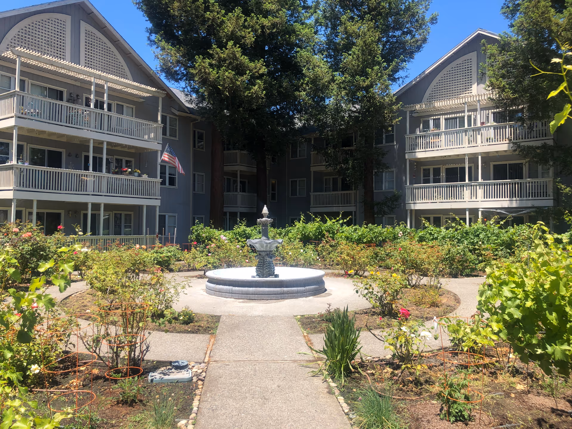 Outdoor courtyard area of a senior living facility with a central water fountain surrounded by garden beds and pathways. The building in the background has multiple balconies and is painted gray. There are trees providing shade and an American flag is visible on one balcony.