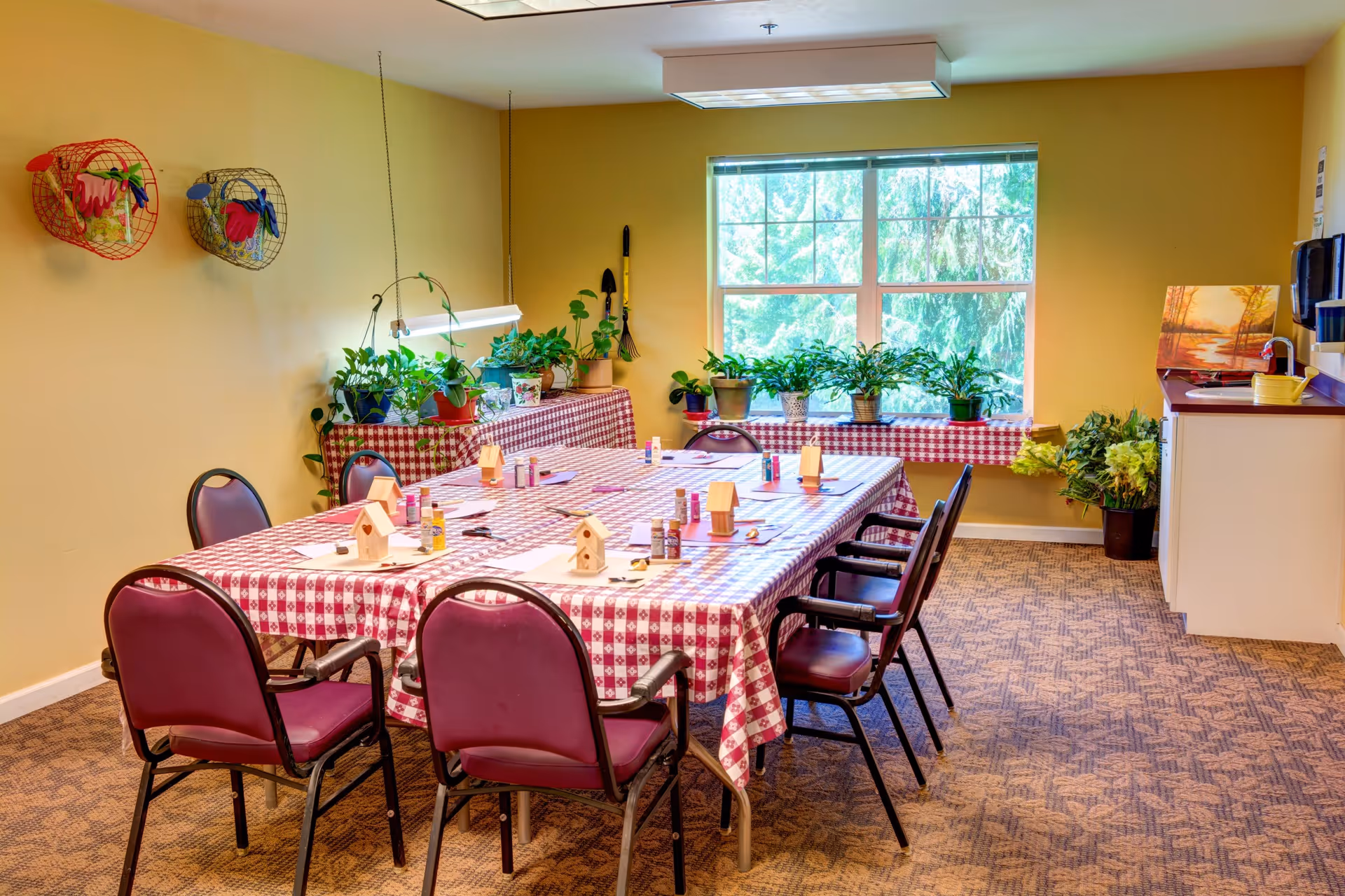 A bright activity room with a large table covered in a red and white checkered tablecloth. The table is set up for a craft activity with small wooden birdhouses, paint bottles, and brushes. Surrounding the table are maroon chairs. The room has yellow walls, a window with a view of greenery outside, and several potted plants on a side table and windowsill. There is a small kitchenette area with a sink and paper towel dispenser on the right side.