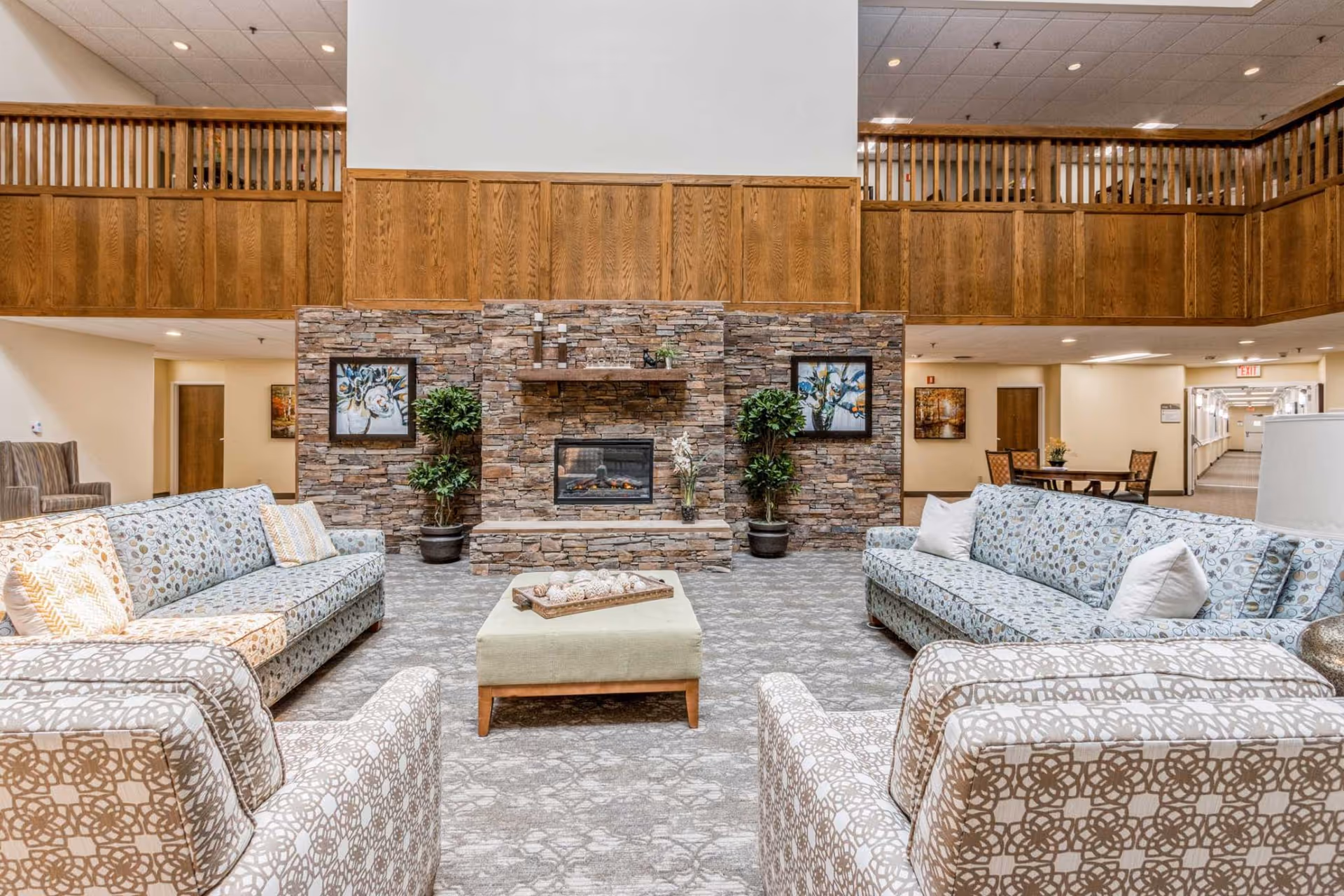 Spacious communal lounge with patterned sofas and chairs arranged around a central ottoman facing a stone fireplace beneath a wooden mezzanine.