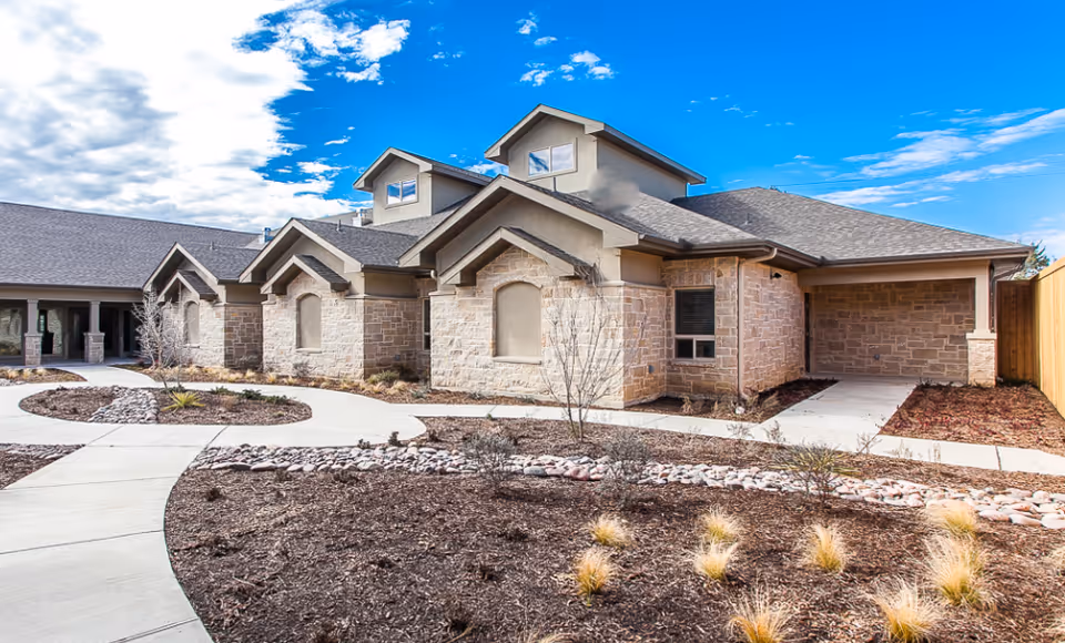 Exterior view of a single-story senior living facility building with stone and stucco walls, multiple gabled roofs, and a clear blue sky with some clouds. The foreground features landscaped garden beds with mulch, rocks, and small plants, along with concrete walkways leading to the building entrance.
