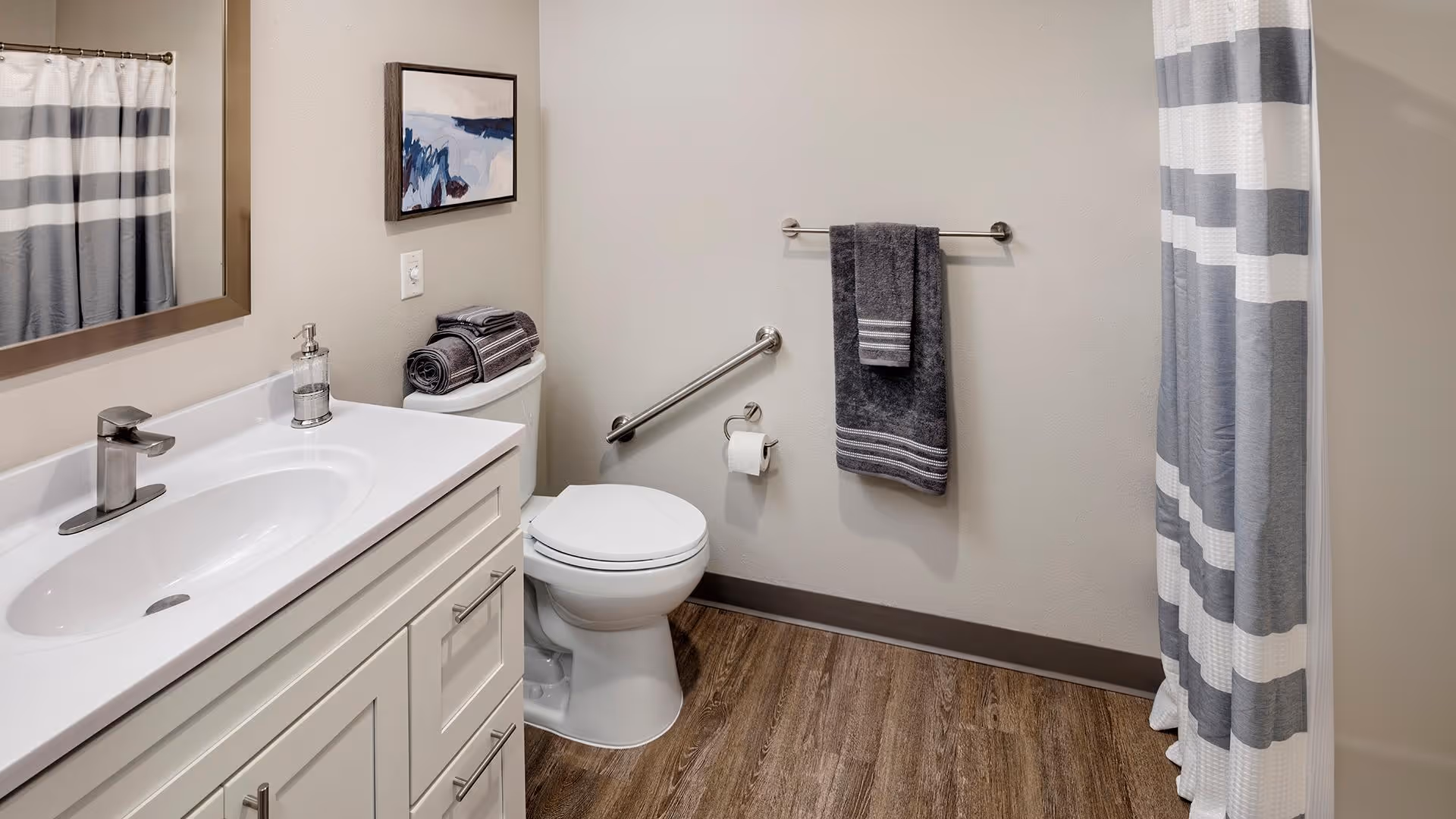A clean and modern bathroom featuring a white vanity with a sink and faucet, a large mirror above it, a toilet with folded towels on top, a towel rack with a dark gray towel, a toilet paper holder, a grab bar on the wall, and a shower with a gray and white striped curtain. The floor has wood-like flooring and the walls are painted light beige.