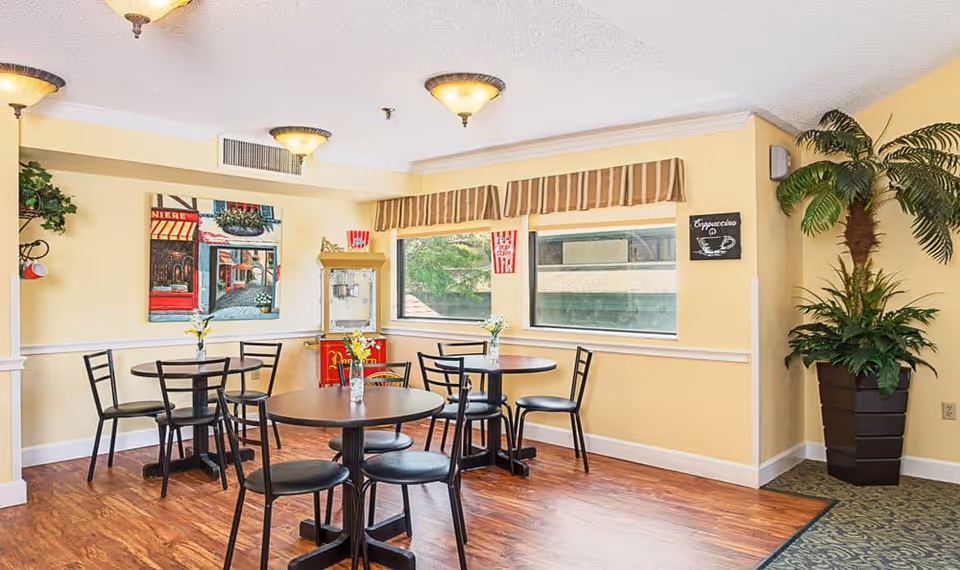 A bright dining area with three round tables, each surrounded by black chairs. The walls are painted yellow and decorated with a colorful painting and a small chalkboard sign that says 'Cappuccino'. There is a popcorn machine in the corner near two large windows with striped valances. A large potted plant is placed in the corner on the right side of the room.