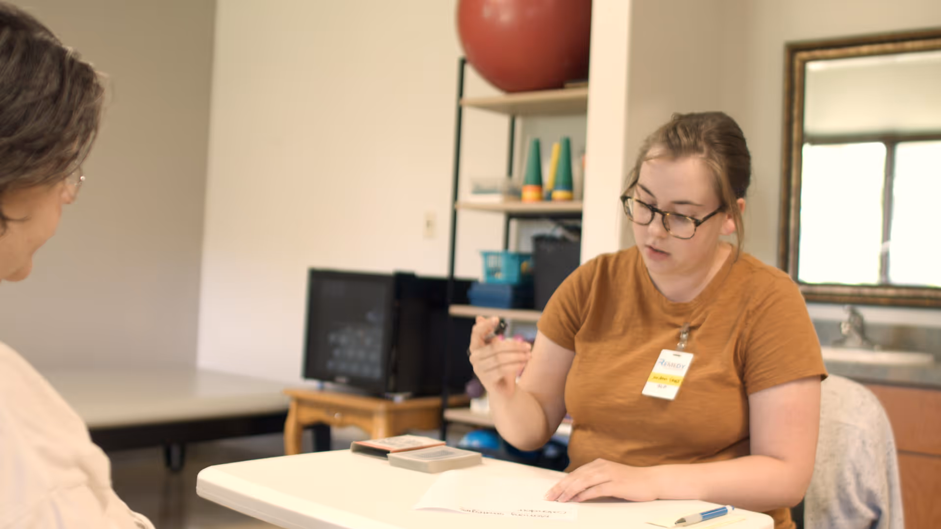 A woman wearing glasses and a brown shirt with a name tag is sitting at a table, holding a small object and looking down at a piece of paper. Another person is partially visible sitting across from her. The room has a shelf with various items, a mirror, and a sink in the background.