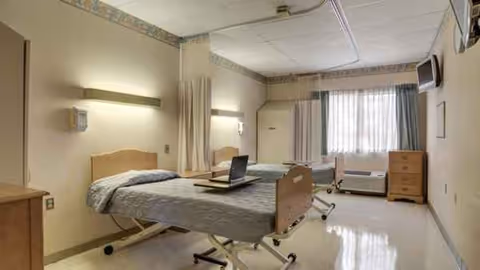 Hospital-style patient room with a bed, laptop, dresser, and windowed curtains.