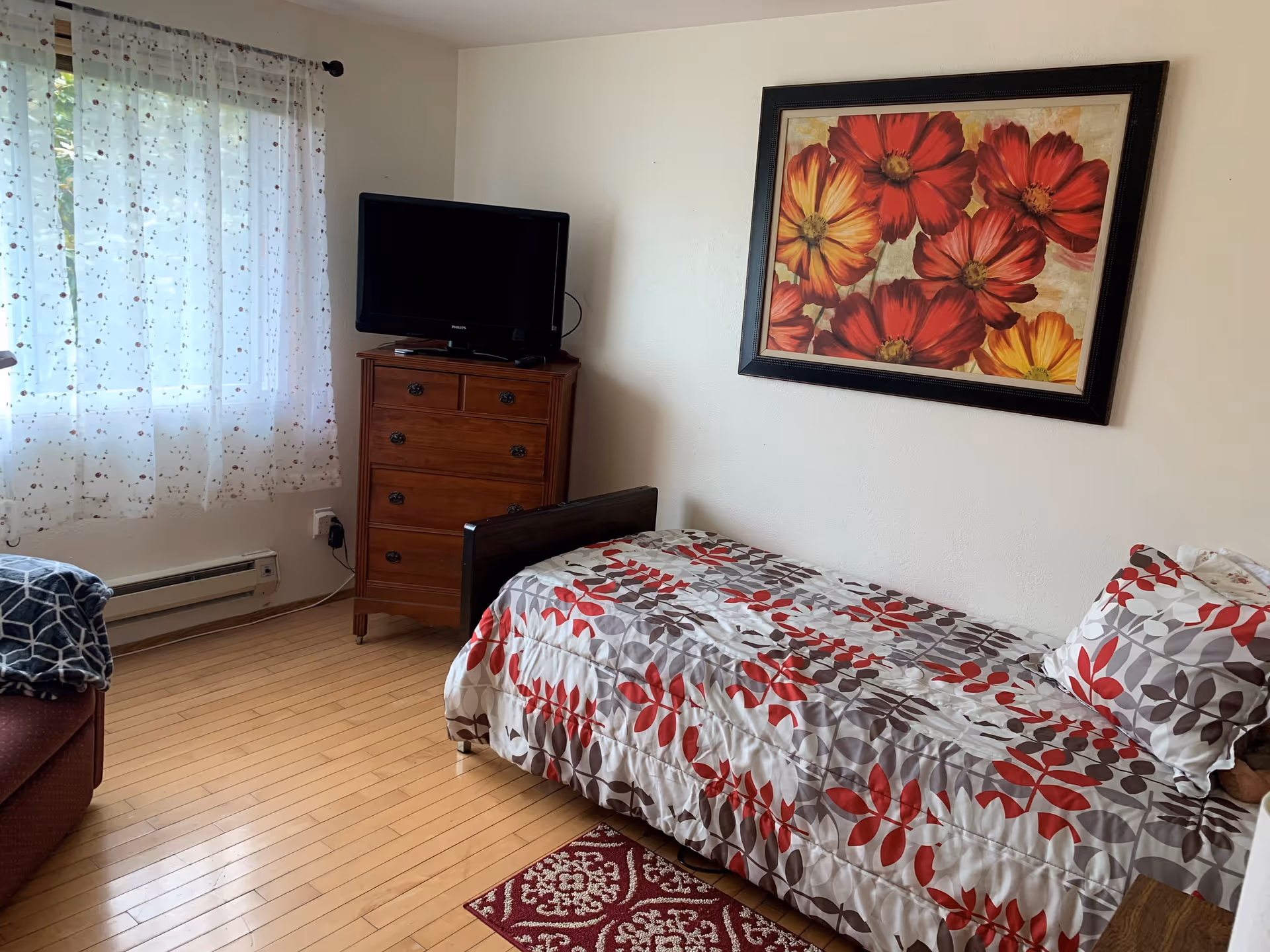 A bedroom with a single bed covered in a white, red, and gray leaf-patterned comforter and matching pillow. Above the bed is a framed painting of large red and yellow flowers. To the left of the bed is a wooden chest of drawers with a flat-screen TV on top. A window with sheer white curtains featuring a small leaf pattern lets in natural light. The floor is light-colored wood with a small red patterned rug partially visible near the bed.
