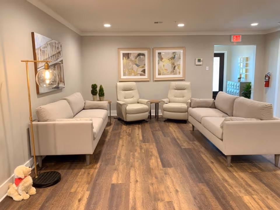 A cozy living room with two beige sofas facing each other and two beige recliner chairs in the middle against a light gray wall. There are two abstract paintings hanging on the wall above the recliners. A modern floor lamp with a round glass shade stands next to one sofa, and a small stuffed dog toy with a red bandana is on the floor nearby. The floor is wooden with a warm tone, and there is a small table between the recliners. A hallway with an exit sign and a fire extinguisher is visible in the background.