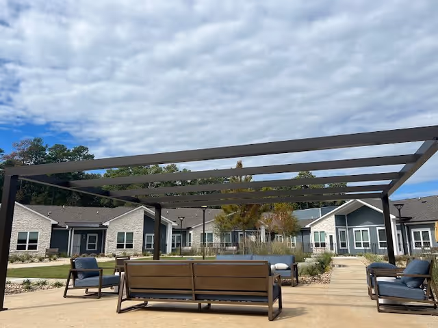 Outdoor courtyard with a pergola and cushioned benches facing single-story residential buildings under a partly cloudy sky.