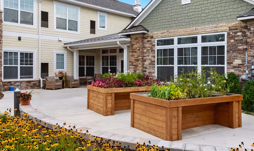 Outdoor patio area at American House Somerset featuring two large wooden raised garden beds filled with various plants and flowers, surrounded by a concrete walkway. The building exterior has beige siding with stone accents and multiple windows with white blinds. There are wicker chairs arranged near the building entrance.