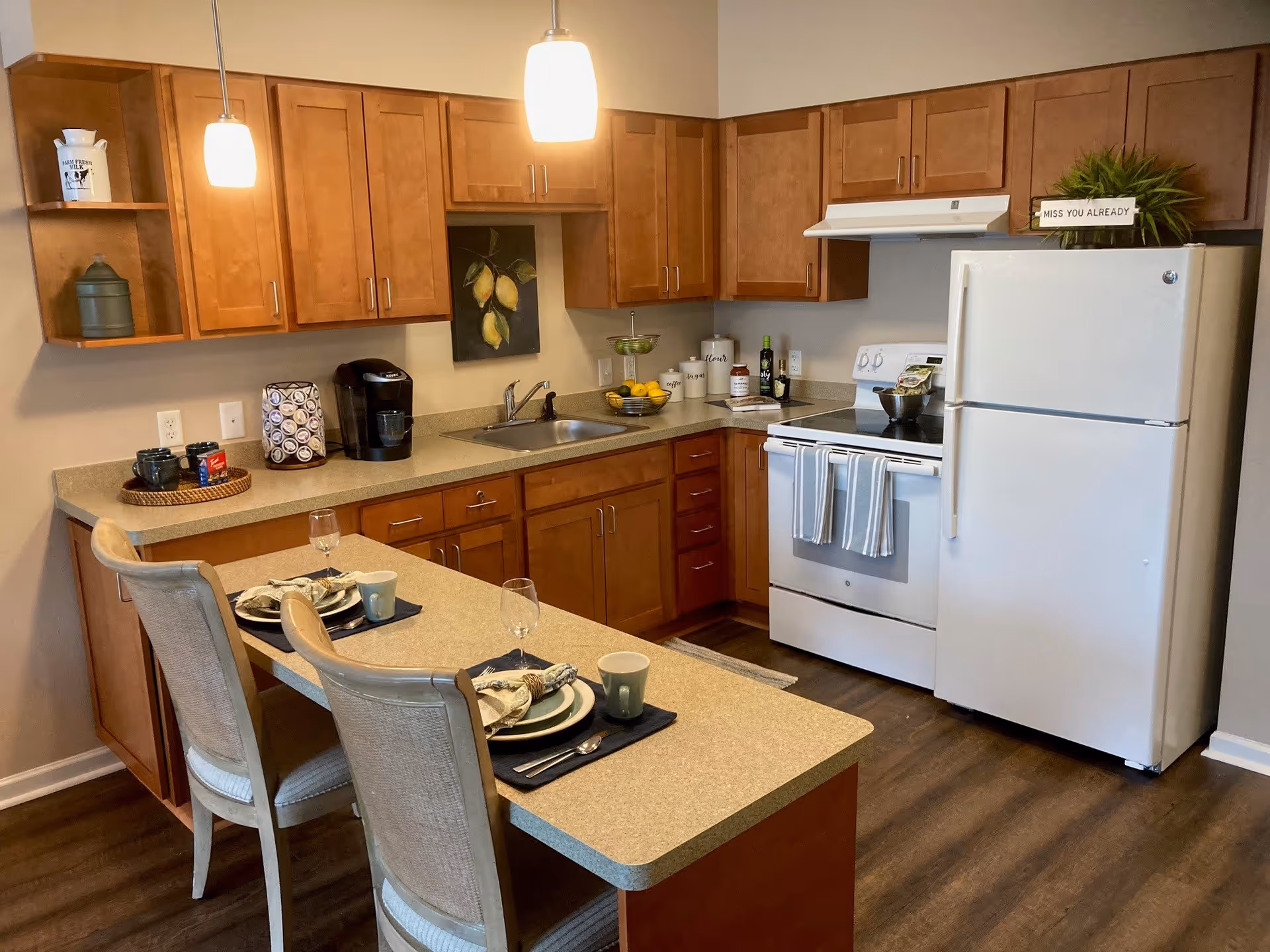 A modern kitchen with wooden cabinets, a white refrigerator, and a white stove with two striped towels hanging on the handle. The kitchen features a countertop with two place settings including plates, napkins, mugs, and wine glasses. There are two pendant lights hanging above the counter, a coffee maker, a fruit bowl with lemons, and decorative items on the shelves and countertop.