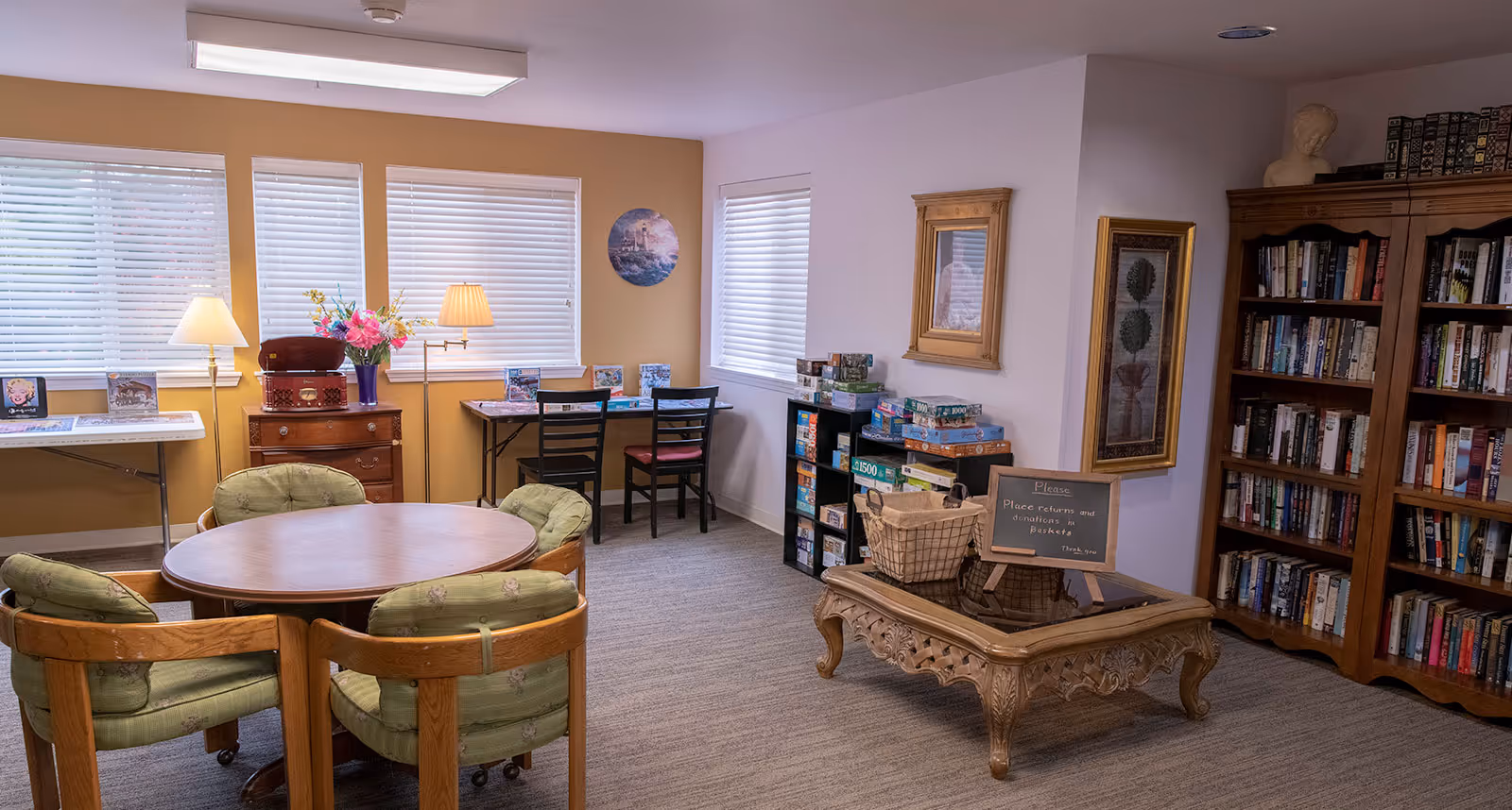 A cozy common room with a round wooden table surrounded by four cushioned chairs, a wooden bookshelf filled with books, a coffee table with a basket and a small chalkboard sign, and two tables near windows with chairs and puzzles on them. The room has beige and white walls with framed artwork and blinds covering the windows.