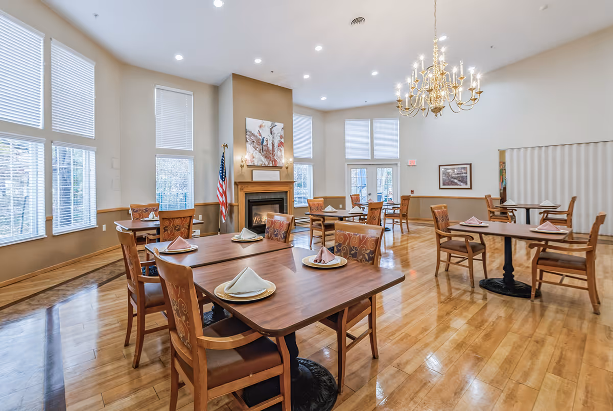 A spacious dining room with several wooden tables and chairs, each table set with plates and folded napkins. The room features large windows with blinds, a fireplace with artwork above it, an American flag, and a chandelier hanging from the ceiling. The floor is polished wood, and the walls are painted in neutral tones.