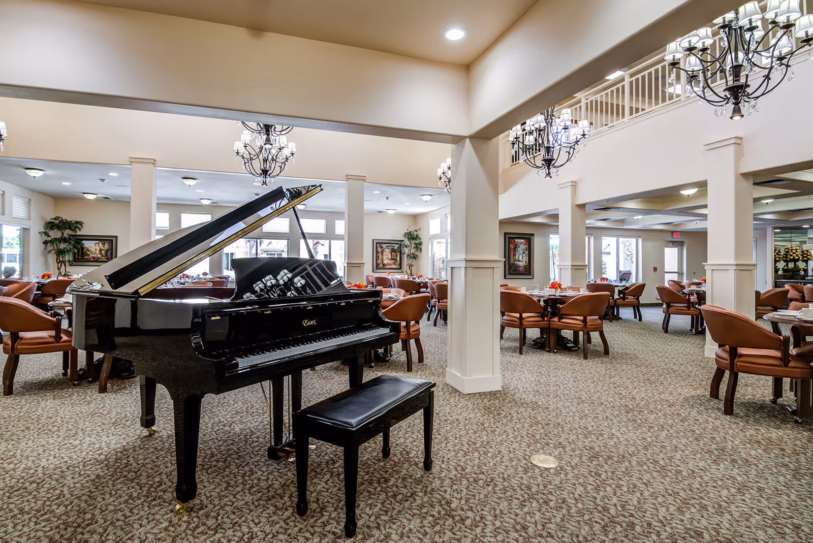 Spacious senior living dining area with a black grand piano and matching bench in the foreground. The room features multiple round tables with brown cushioned chairs, chandeliers hanging from the ceiling, large windows allowing natural light, and decorative plants and paintings on the walls.