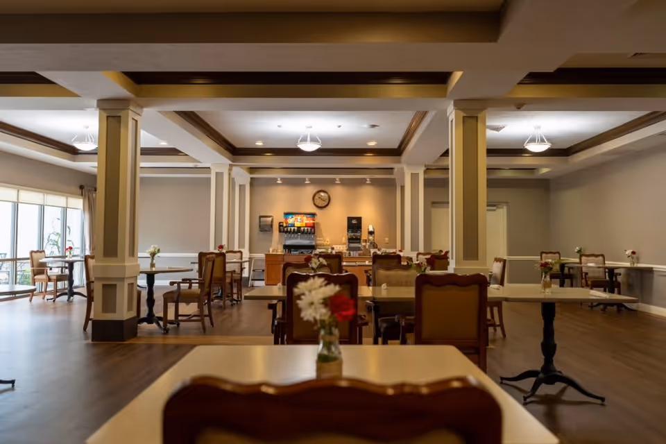 A spacious dining room with multiple tables and chairs arranged neatly. Each table has a small vase with flowers. The room features wooden flooring, white pillars, and a ceiling with recessed lighting and decorative beams. At the far end, there is a beverage station with a soda dispenser and coffee machine beneath a wall clock.