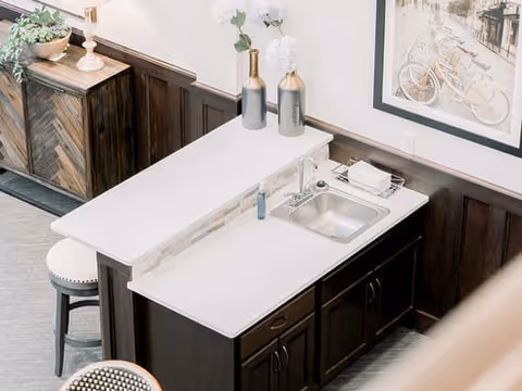 A small kitchenette area with a white countertop, a stainless steel sink, two decorative vases with white flowers, and a soap dispenser. There is a wooden cabinet below the counter and a wooden sideboard with a plant and a lamp in the background. A framed picture of a bicycle is hanging on the wall above the counter.