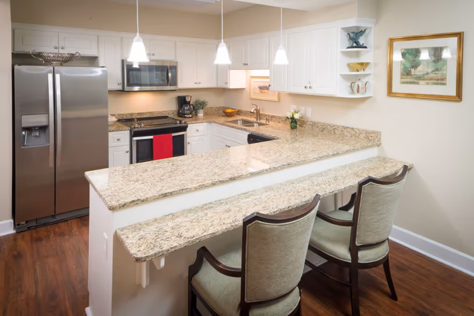 Modern kitchen with granite countertops, white cabinets, stainless steel refrigerator and microwave, a stove with a red towel hanging on the handle, two upholstered chairs at a breakfast bar, pendant lights hanging from the ceiling, and framed artwork on the wall.