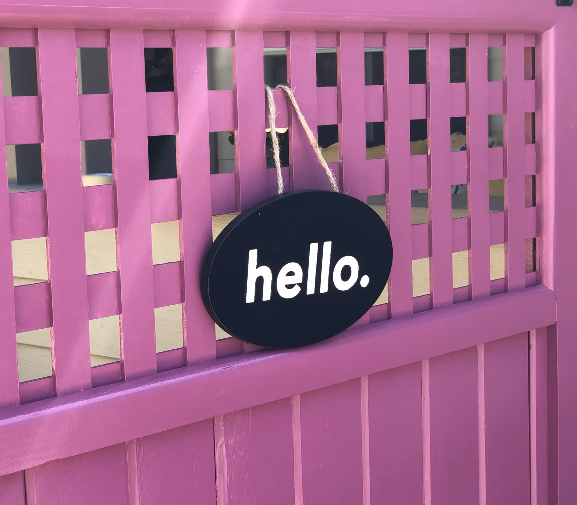 A black oval sign with the word 'hello.' in white letters hanging on a purple wooden lattice fence with a twine string.