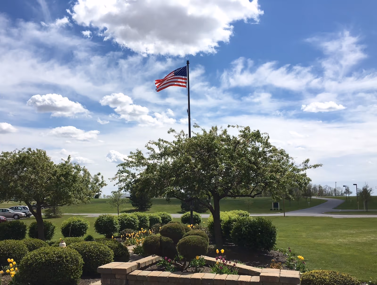 A landscaped outdoor garden area with neatly trimmed bushes, flowering plants including yellow and purple tulips, and two small trees. An American flag on a tall flagpole is waving in the breeze against a partly cloudy blue sky. A paved road and parking area are visible in the background.