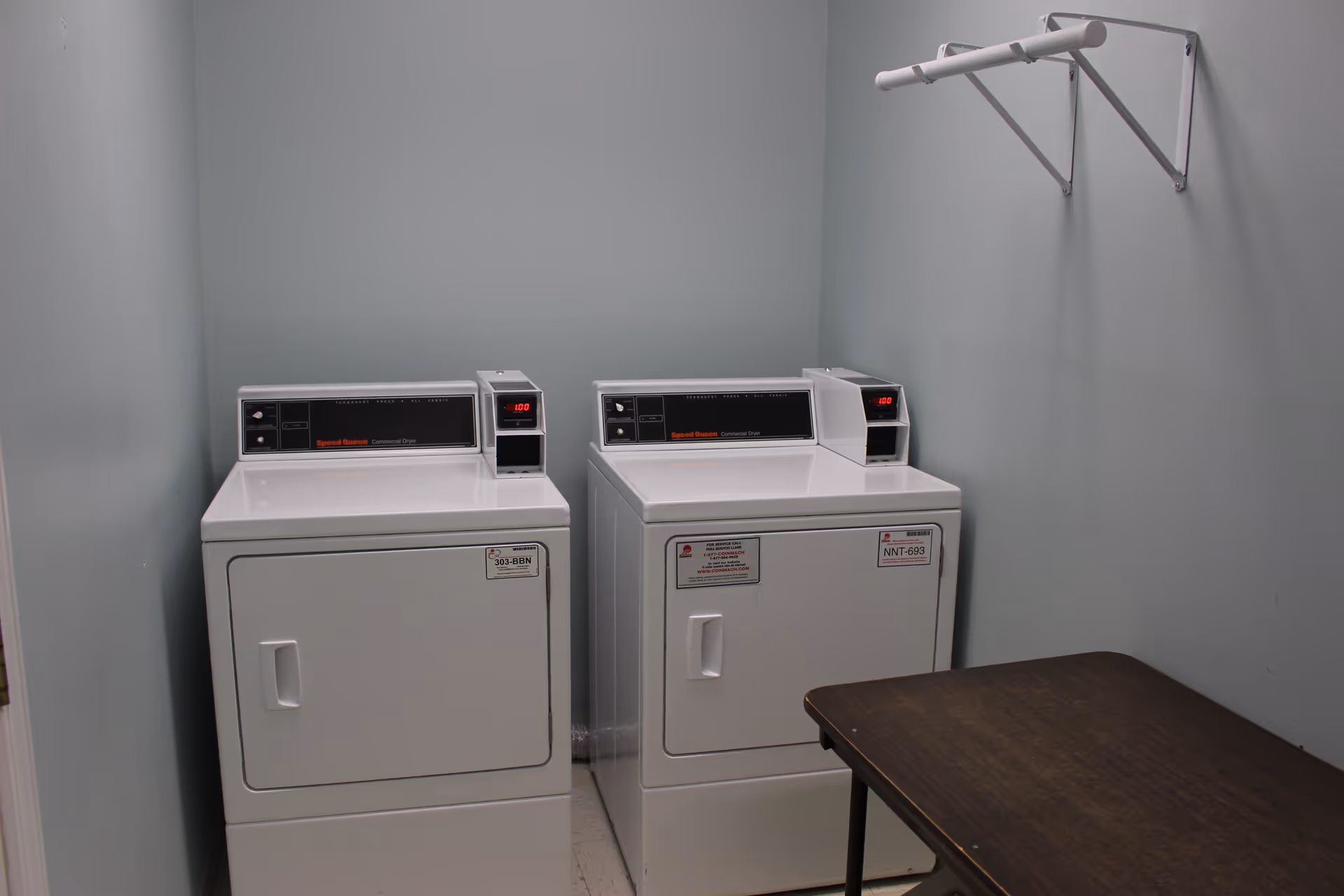 A small laundry room with two white commercial Speed Queen dryers side by side against a light gray wall. Above the dryers is a white wall-mounted drying rack. A dark brown folding table is positioned to the right of the dryers.