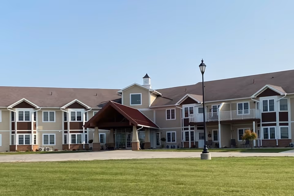 Exterior view of a two-story senior living facility building with beige and brown siding, multiple windows, a covered entrance with a red roof, and a lamp post in front. The building is surrounded by a well-maintained green lawn under a clear blue sky.