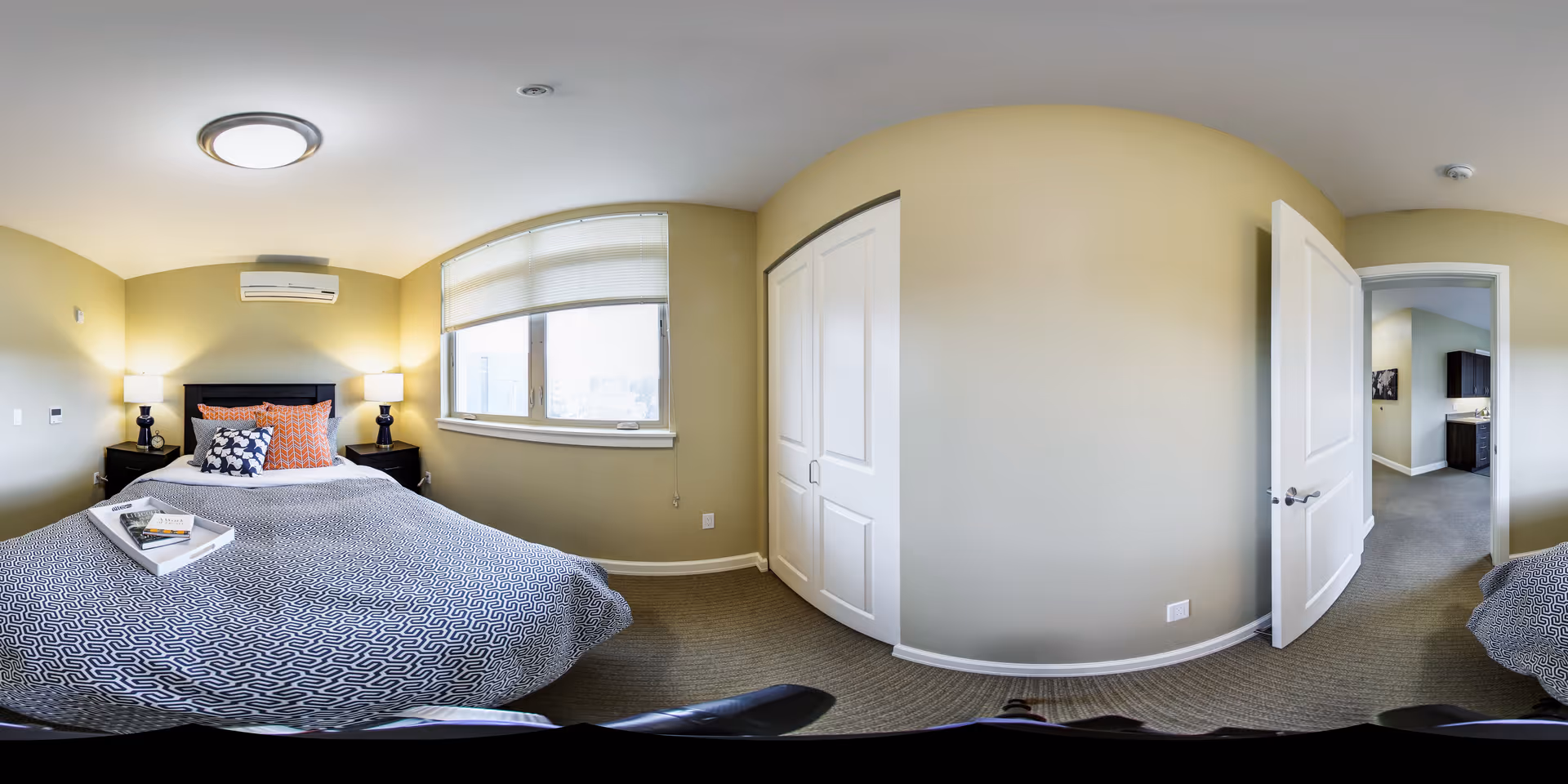 A well-lit bedroom with a large bed covered in a patterned gray and white bedspread, two bedside tables with lamps, a window with blinds, a closet with double doors, and an open door leading to another room.