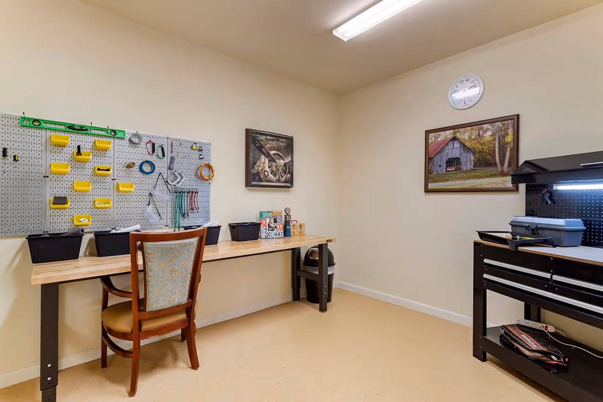A small workshop or craft room with a wooden table and a chair. On the wall above the table is a pegboard with various tools and supplies organized in yellow holders. There are two framed pictures on the walls, one showing a barn and the other a close-up of a mechanical object. A clock is mounted on the wall above the barn picture. To the right, there is a black tool chest with additional tools and supplies. The room has beige walls and a beige floor with overhead fluorescent lighting.