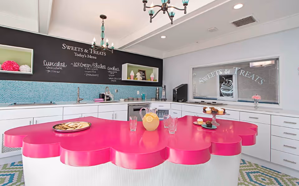 Bright and colorful kitchen area with a large pink scalloped island countertop. The back wall features a blackboard menu titled 'Sweets & Treats' listing cupcakes, ice cream shakes, and cookies. The kitchen has white cabinets, a sink, and a window with 'Sweets & Treats' and a cupcake design etched on the glass. On the island, there is a pitcher of lemonade, glasses, a plate of cookies, and a tiered tray with cupcakes.