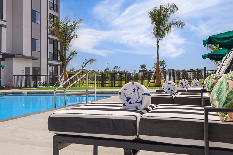 Outdoor pool area with striped lounge chairs, each topped with a rolled towel, palm trees, and a clear blue sky. A multi-story building is visible on the left side.