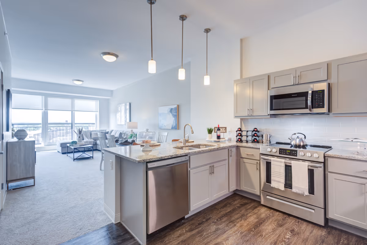 Modern open-concept kitchen and living room area with light gray cabinets, stainless steel appliances including a microwave, stove, and dishwasher. The kitchen island has a sink and pendant lights hanging above. The living room features a large window with blinds, a sofa, coffee table, and wall art.