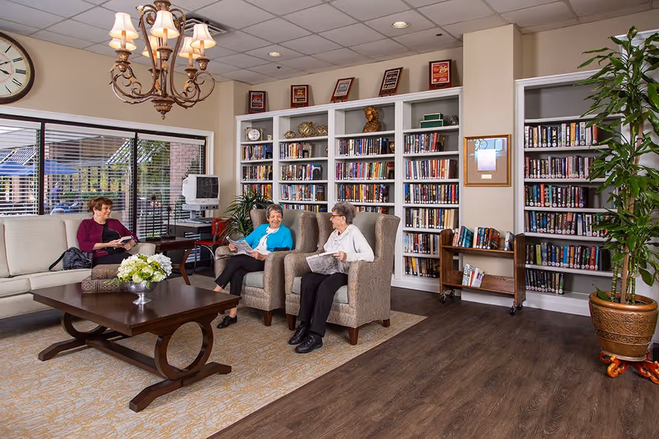 A cozy library or common area in a retirement community with three elderly women sitting and reading newspapers and books. The room features a large wooden coffee table with a flower arrangement, comfortable armchairs and a sofa, a bookshelf filled with books, a large clock on the wall, a chandelier, and a potted plant.