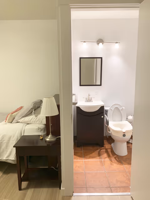 View of a bedroom and an adjacent bathroom. The bedroom has a bed with pillows and a nightstand with a lamp. The bathroom features a toilet with armrests, a sink with a dark cabinet, a mirror above the sink, and a light fixture with three bulbs on the wall.