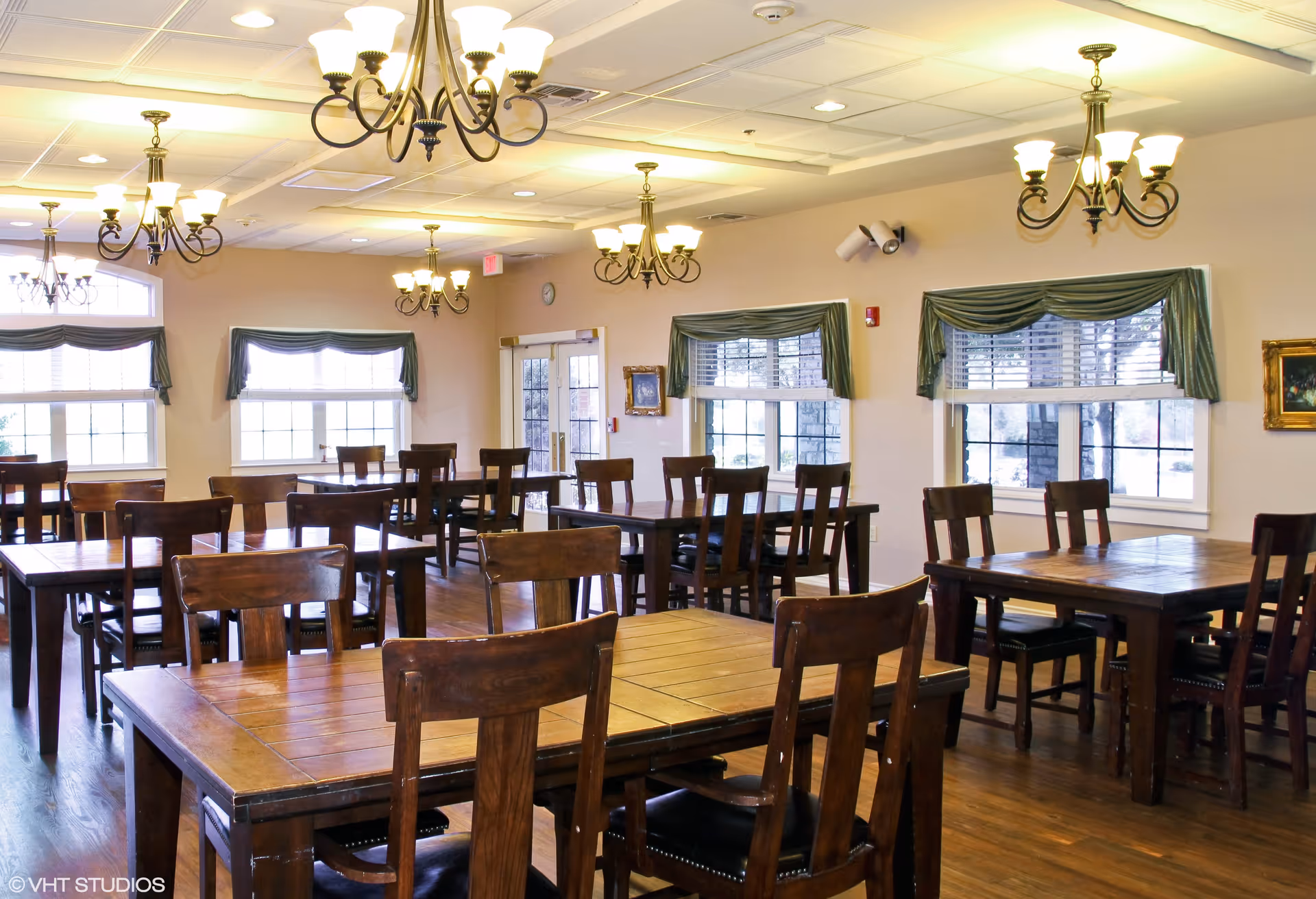 A bright dining room with multiple wooden tables and chairs arranged neatly. The room features large windows with green valances, wooden flooring, and several ornate chandeliers hanging from the ceiling. The walls are light-colored, and there is a door at the far end of the room.