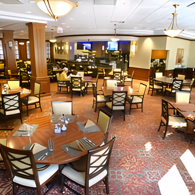 A spacious dining room with multiple round and square tables, each surrounded by cushioned chairs. The tables are set with placemats, napkins, and glassware. The room features warm lighting, carpeted floors with a patterned design, and wood-paneled columns and walls. There are large windows and a mirror reflecting the room, enhancing the sense of space.
