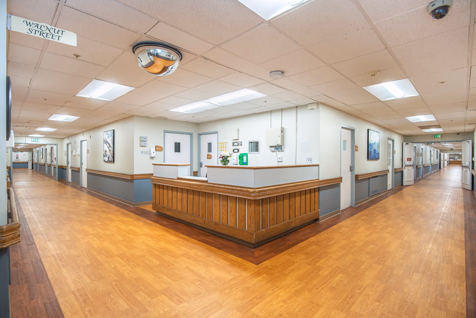Wide, well-lit interior hallway with a central nurses' station, wood-accented floors and doors lining both sides.