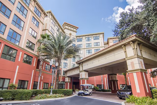 Exterior view of a multi-story senior living facility with a covered entrance, palm trees, and parked vehicles including a golf cart and a car under the canopy.