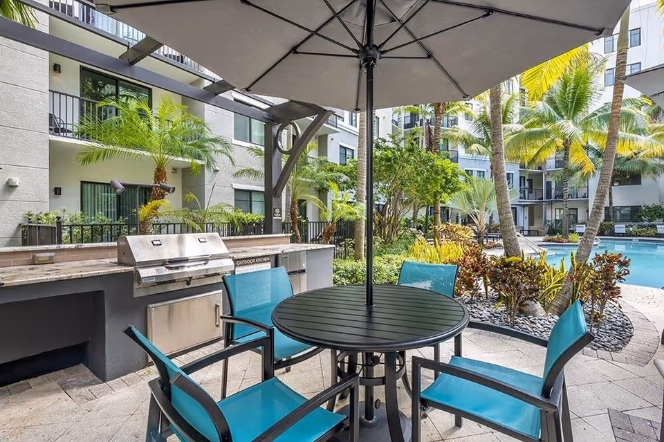 Poolside outdoor seating area with an umbrella-covered round table, turquoise chairs, a built-in grill, palm trees and apartment balconies in the background.