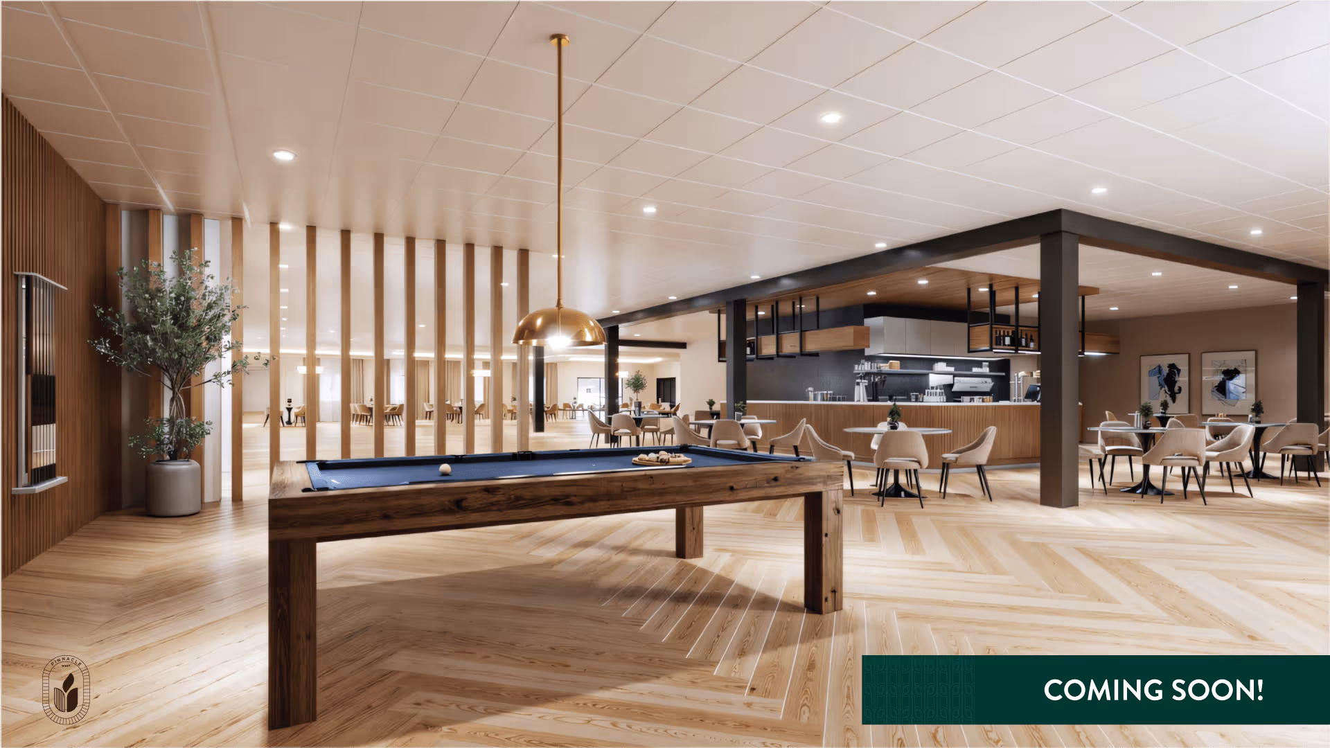 A spacious and modern common area featuring a wooden pool table with a blue felt top in the foreground. Behind the pool table, there are multiple round tables with beige chairs arranged for seating. The area includes a coffee bar or kitchen counter with hanging shelves and modern lighting. The floor is light wood with a herringbone pattern, and the ceiling has recessed lighting. A large plant in a pot is placed near a wooden slatted partition wall on the left side. The bottom left corner shows a Pinnacle West logo, and the bottom right corner has a green banner with the text 'COMING SOON!'