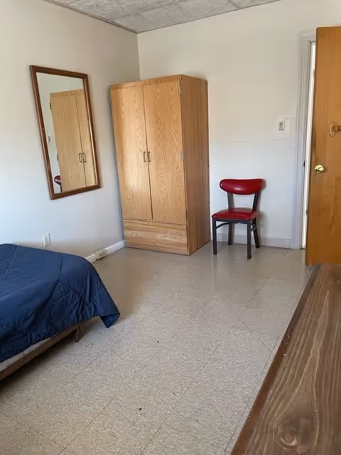 A simple bedroom with a bed covered in a dark blue blanket, a wooden wardrobe, a red chair, a wall-mounted mirror, and a wooden door. The floor is tiled and the walls are plain white.