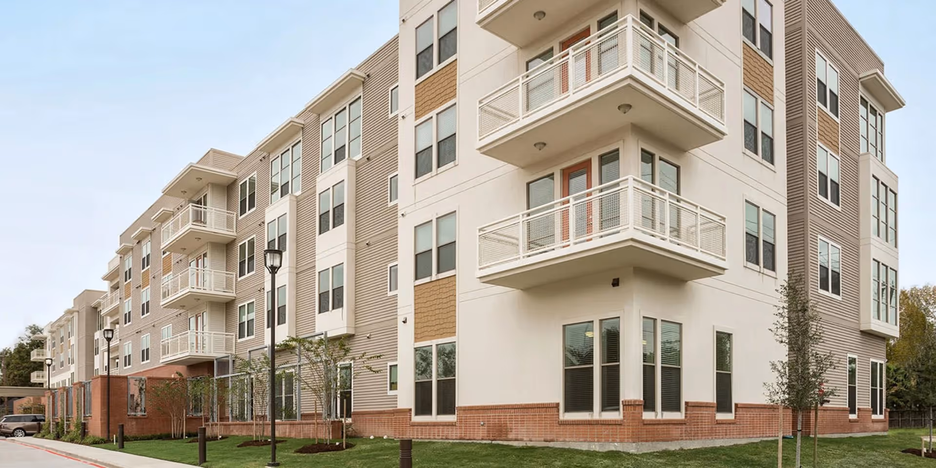 Modern multi-story senior living apartment building with balconies and small landscaped grounds under a pale sky.