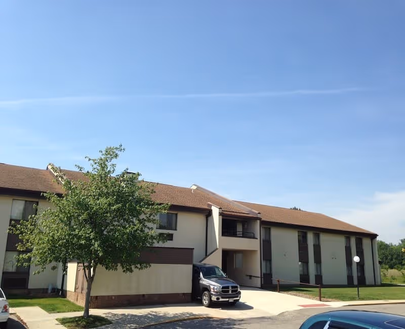 Exterior view of a two-story senior living facility building with beige walls and a brown roof under a clear blue sky. There is a tree in front of the building and several parked vehicles in the parking area.