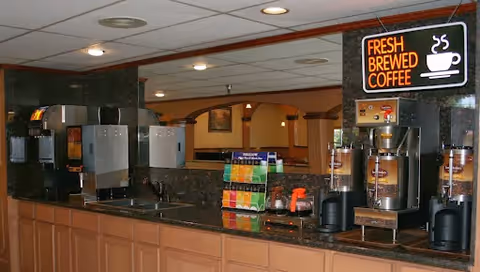 Interior view of a coffee and beverage station with multiple coffee dispensers, a soda fountain machine, and a sign that reads 'Fresh Brewed Coffee' in a senior living facility.