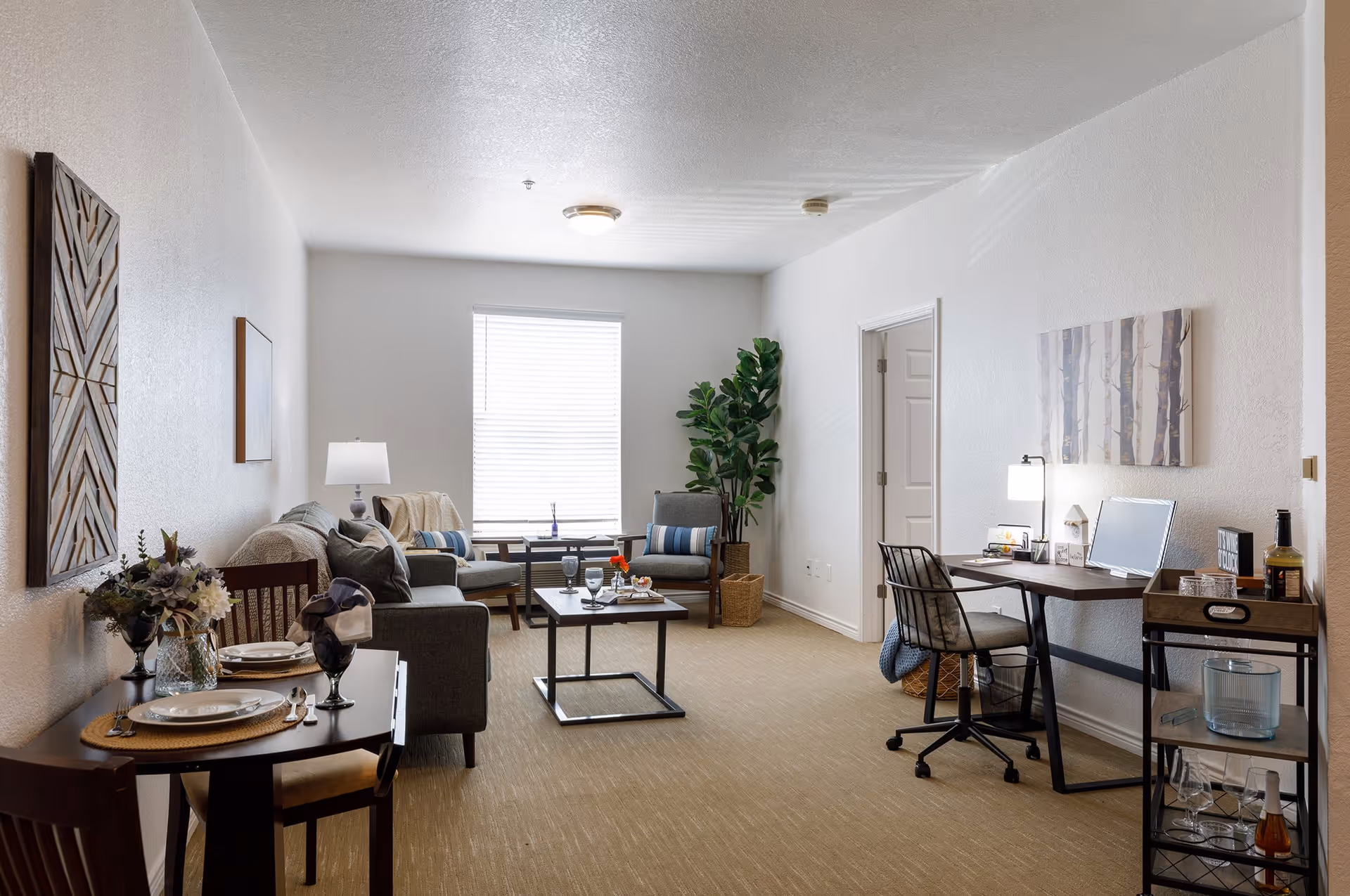A bright and neatly arranged living room in a senior living facility with a small dining table set for two on the left, a gray sofa and armchair near a window with blinds, a coffee table with glasses and a flower vase, a potted plant in the corner, and a desk with a chair, lamp, and computer on the right side. The walls are decorated with framed artwork.