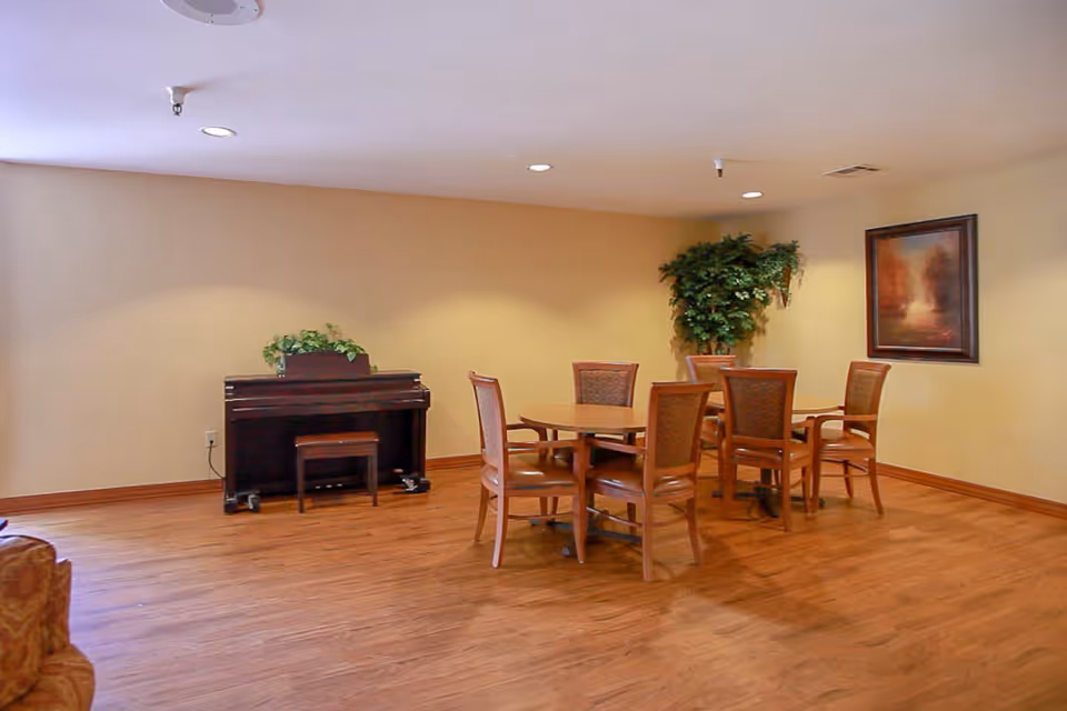 A room with wooden flooring featuring a round wooden table surrounded by six chairs. Against the wall is a dark wooden piano with a small bench and a green plant on top. There is a large potted plant in the corner and a framed painting on the wall. The walls are painted beige and the ceiling has recessed lighting.