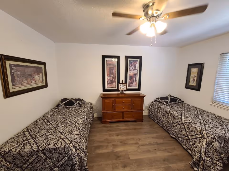 A bedroom with two twin beds on opposite sides of the room, each covered with a black and white patterned bedspread. Between the beds is a wooden dresser with three drawers and two cabinet doors. Above the dresser are two framed vertical pictures and a small framed photo on a stand. The walls are white, and there are three framed pictures hanging on the walls. A ceiling fan with lights is on the ceiling, and a window with blinds is on the right wall.