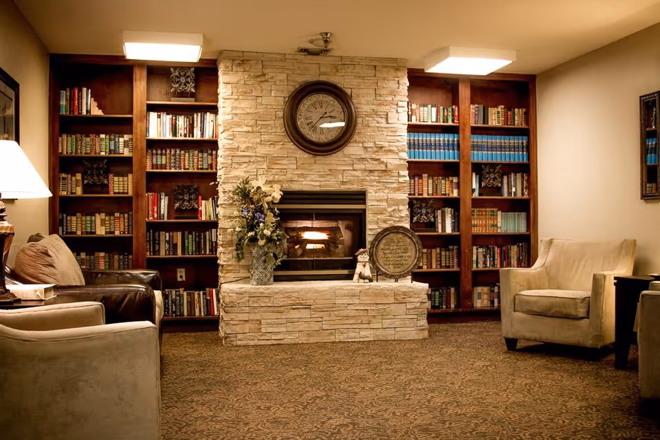 Cozy living room area with a stone fireplace in the center, a round clock above it, and built-in wooden bookshelves filled with books on either side. There are two armchairs, one beige and one brown leather, positioned around the fireplace, along with a table lamp on the left side. The carpet has a patterned design and the room is warmly lit.